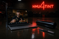 HMU4TINT Install Confidence Credit — laptop on wet concrete showing a tint installer on a live video call, with tint film and knife nearby under HMU4TINT neon